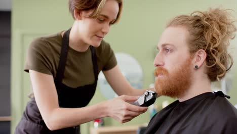 Happy-caucasian-female-barber-trimming-beard-of-red-haired-male-client-at-barbershop,-in-slow-motion