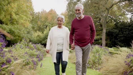 Senior-biracial-couple-holding-hands-and-walking-together-in-sunny-garden,-unaltered,-in-slow-motion