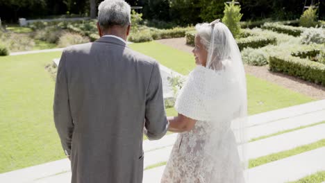 Happy-senior-biracial-couple-walking,-holding-hands-at-wedding-in-garden,-slow-motion