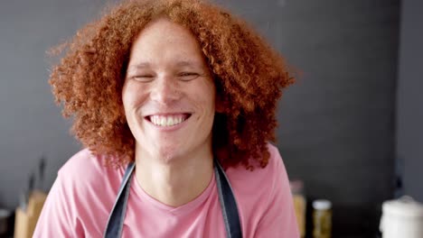 Portrait-of-happy-biracial-man-with-red-curly-hair-wearing-apron-smiling-in-kitchen,-in-slow-motion