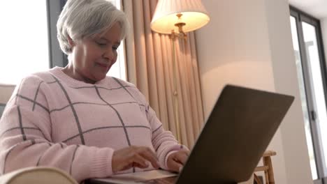 Glückliche-ältere-Biraziale-Frau,-Die-Im-Sessel-Sitzt-Und-Einen-Laptop-Benutzt,-Unverändert,-In-Zeitlupe