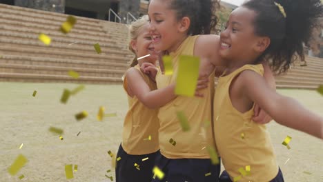 Animation-of-gold-confetti-over-happy-diverse-schoolgirls-celebrating-and-hugging-on-sports-field