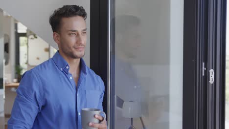 Happy-biracial-man-holding-holding-coffee-looking-through-window-in-modern-apartment,-slow-motion