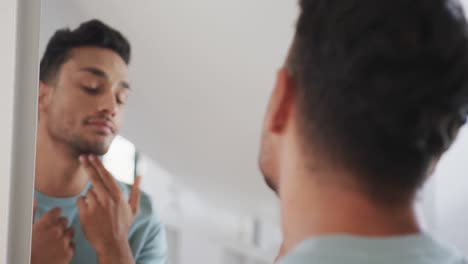 Happy-biracial-man-looking-in-bathroom-mirror,-examining-his-face-and-skin,-slow-motion