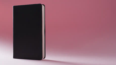 Close-up-of-closed-black-book-standing-vertical-with-copy-space-on-pink-background-in-slow-motion