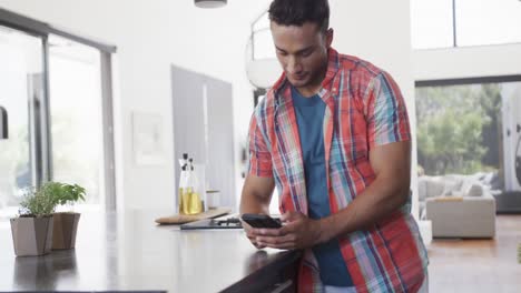 Happy-biracial-man-leaning-on-countertop-in-sunny-kitchen-using-smartphone,-slow-motion