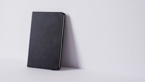 Close-up-of-closed-black-book-leaning-on-wall-with-copy-space-on-white-background-in-slow-motion