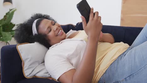Happy-african-american-plus-size-woman-lying-on-sofa-and-using-smartphone,-unaltered,-in-slow-motion