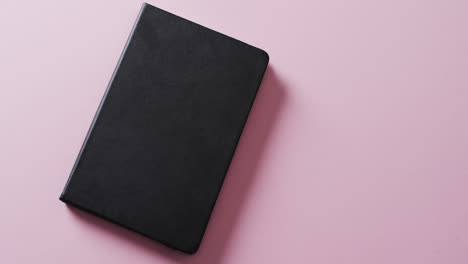 Close-up-of-closed-black-book-with-copy-space-on-pink-background-in-slow-motion