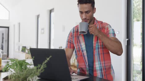 Biracial-man-having-coffee-and-using-laptop-in-sunny-modern-apartment,-slow-motion