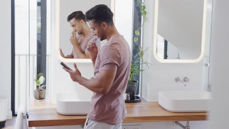 Biracial-man-brushing-teeth-in-the-morning-and-using-smartphone-in-sunny-bathroom,-slow-motion