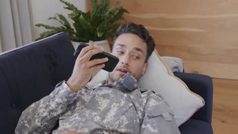 Happy-biracial-male-soldier-in-uniform-lying-on-sofa-at-home-talking-on-smartphone,-slow-motion