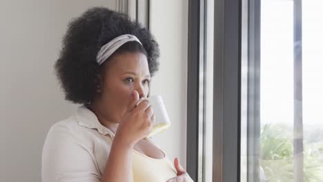 African-american-plus-size-woman-looking-through-window-and-holding-mug,-unaltered,-slow-motion