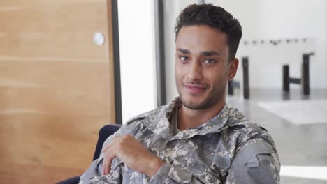 Portrait-of-happy-biracial-male-soldier-in-uniform-sitting-in-armchair-at-home-smiling,-slow-motion