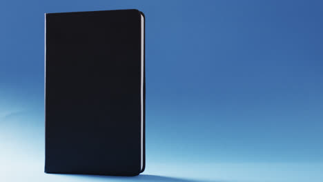 Close-up-of-closed-black-book-standing-vertical-with-copy-space-on-blue-background-in-slow-motion