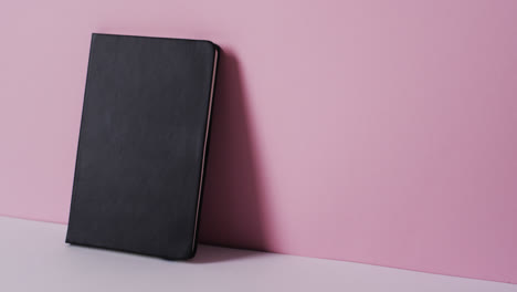 Close-up-of-closed-black-book-leaning-on-wall-with-copy-space-on-pink-background-in-slow-motion