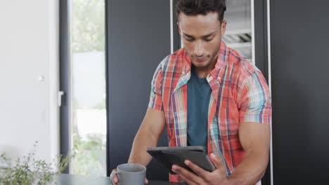 Biracial-man-having-coffee-and-using-tablet-in-kitchen-at-home,-slow-motion