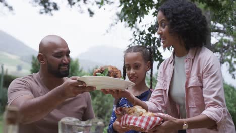 Happy-biracial-parents-and-daughter-serving-meal-at-dinner-table-in-garden,-slow-motion