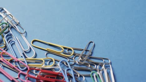 Close-up-of-scattered-colourful-paper-clips-with-copy-space-on-blue-background,-in-slow-motion