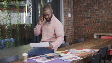 Busy-african-american-casual-businessman-sitting-on-desk,-calling-and-smiling