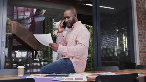 African-american-casual-businessman-with-document-talking-on-smartphone-sitting-on-desk,-slow-motion