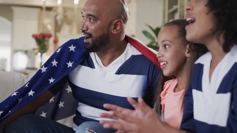 Aufgeregt-Gemischtrassige-Eltern-Und-Tochter,-Die-Zu-Hause-Sport-Im-Fernsehen-Mit-Amerikanischer-Flagge-Schauen,-Zeitlupe