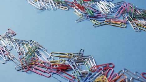 Close-up-of-scattered-colourful-paper-clips-with-copy-space-on-blue-background,-in-slow-motion
