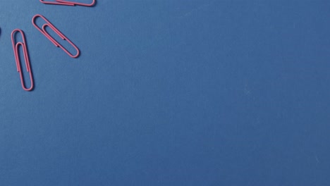 Overhead-view-of-pink-paper-clips-arranged-with-copy-space-on-blue-background,-in-slow-motion