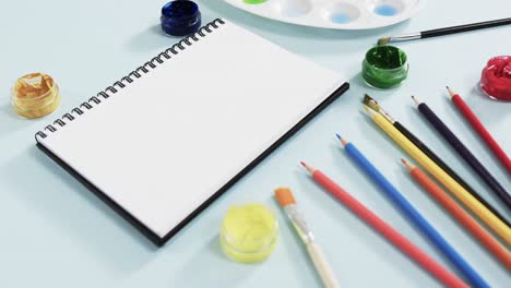 Primer-Plano-De-Cuaderno,-Pinceles-Y-Pinturas-Sobre-Fondo-Azul,-En-Cámara-Lenta