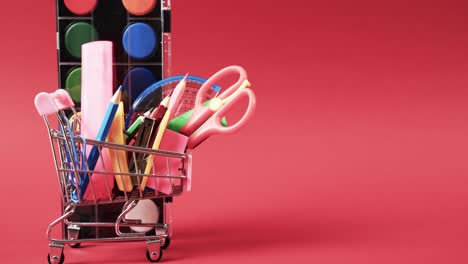Close-up-of-shopping-trolley-with-school-items-and-paints-with-copy-space-on-red-background