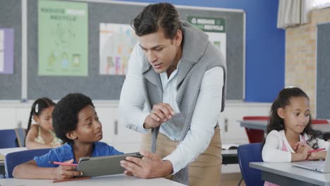 Happy-diverse-male-teacher-helping-schoolchildren-using-tablet-in-classroom-at-elementary-school