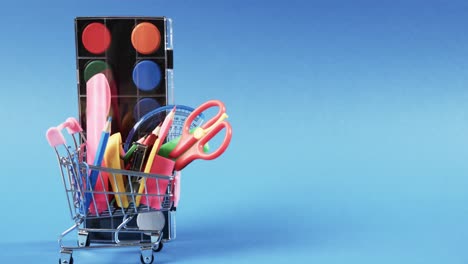 Close-up-of-shopping-trolley-with-school-items-and-paints-with-copy-space-on-blue-background