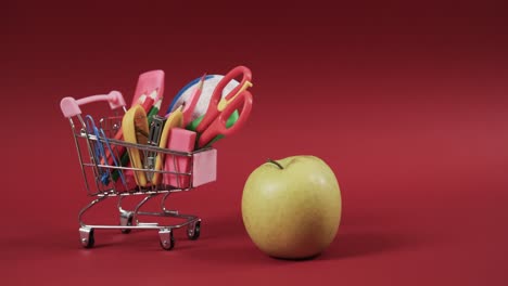 Close-up-of-shopping-trolley-with-school-items-and-apple-with-copy-space-on-red-background
