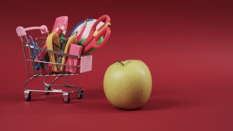 Close-up-of-shopping-trolley-with-school-items-and-apple-with-copy-space-on-red-background
