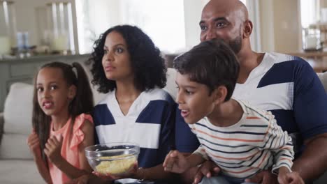 Happy-biracial-parents,-son-and-daughter-watching-sport-on-tv-with-popcorn-at-home,-slow-motion