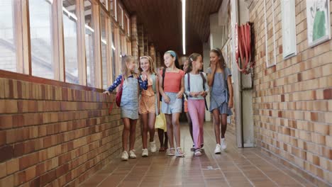 Happy-diverse-schoolgirls-with-school-bags-walking-in-corridor-at-elementary-school