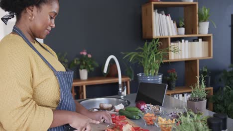 Profile-of-happy-african-american-woman-chopping-vegetables-and-using-tablet-in-kitchen,-slow-motion