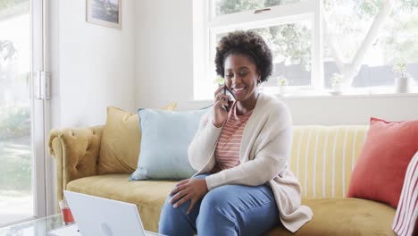 Happy-african-american-woman-working-at-home,-talking-on-smartphone-and-using-laptop,-slow-motion