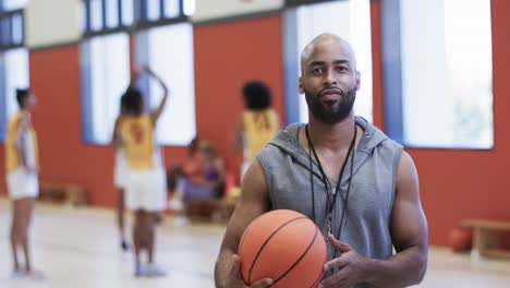 Portrait-of-african-american-male-basketball-coach-holding-ball-in-indoor-court,-in-slow-motion
