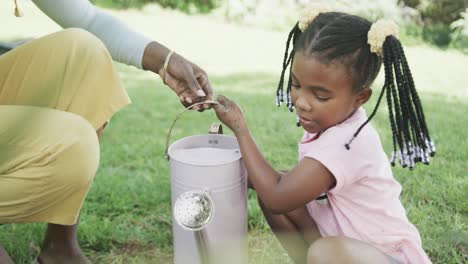 Happy-african-american-mother-and-daughter-watering-flowers-in-sunny-garden,-slow-motion