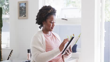 Portrait-of-happy-african-american-woman-using-tablet-standing-in-sunny-kitchen-at-home,-slow-motion