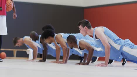 Diverse-male-basketball-coach-watching-team-doing-press-ups-at-indoor-court,-slow-motion