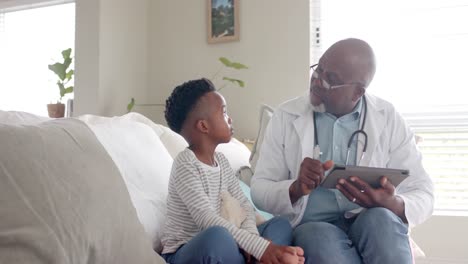 Senior-african-american-male-doctor-using-tablet-talking-to-boy-patient,-slow-motion