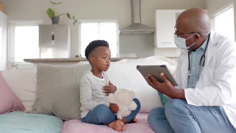 Senior-african-american-male-doctor-in-face-mask-using-tablet-talking-to-boy-patient,-slow-motion