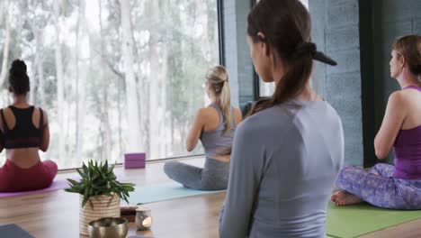 Vista-Trasera-De-Diversas-Mujeres-Practicando-Meditación-Mientras-Están-Sentadas-En-Colchonetas-En-Un-Estudio-De-Yoga.