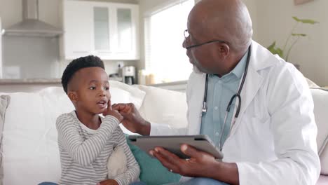 Senior-african-american-male-doctor-using-tablet-talking-to-boy-patient-at-home,-slow-motion