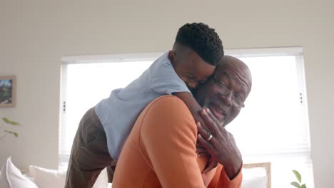 Happy-african-american-grandson-and-grandfather-embracing-and-smiling-on-couch-at-home,-slow-motion