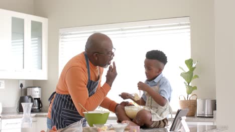 Happy-african-american-grandfather-and-grandson-using-tablet,-baking-in-kitchen,-slow-motion