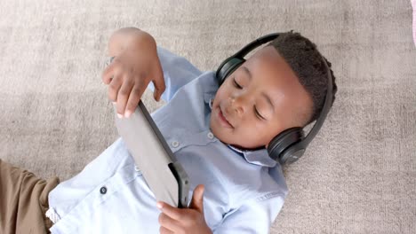 Feliz-Niño-Afroamericano-Con-Auriculares-Tirados-En-El-Suelo,-Usando-Tableta-En-Casa,-Cámara-Lenta
