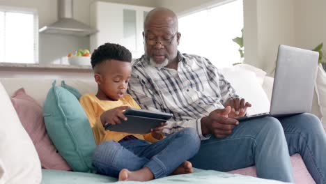 Afroamerikanischer-Großvater-Mit-Enkel-Mit-Tablet-Und-Laptop-Auf-Der-Couch-Zu-Hause,-Zeitlupe